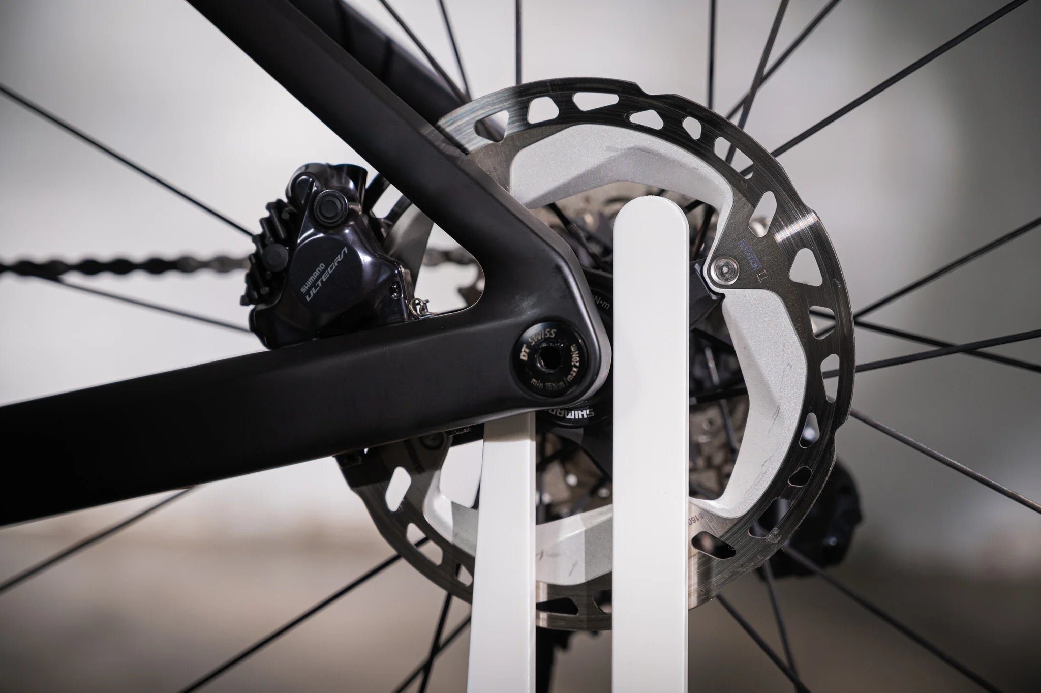 Detailed close-up of the fork-shaped mount on the white Hold My Bike (HMB) bike stand, securely holding the rear wheel via the thru-axle without touching the carbon frame or brake rotor.