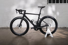 A matte black Rose XLITE 06 road bike displayed in the minimalist white Hold My Bike (HMB) bike stand against a light brick wall.