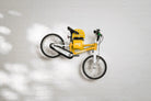 A yellow Hold My Bike (HMB) Kids wall mount securely holding a yellow woom 2 kids bike by the seat post, with a matching yellow woom helmet resting on the integrated top shelf.