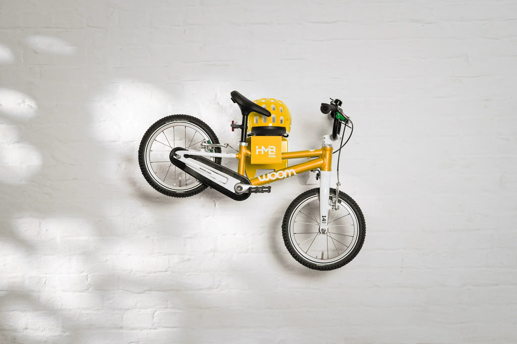 A yellow Hold My Bike (HMB) Kids wall mount securely holding a yellow woom 2 kids bike by the seat post, with a matching yellow woom helmet resting on the integrated top shelf.