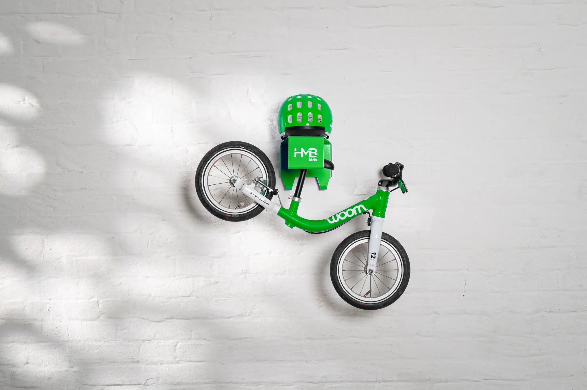 A green Hold My Bike (HMB) Kids wall mount securely holding a green woom 1 balance bike by the seat post, with a matching green woom helmet resting on the integrated top shelf.