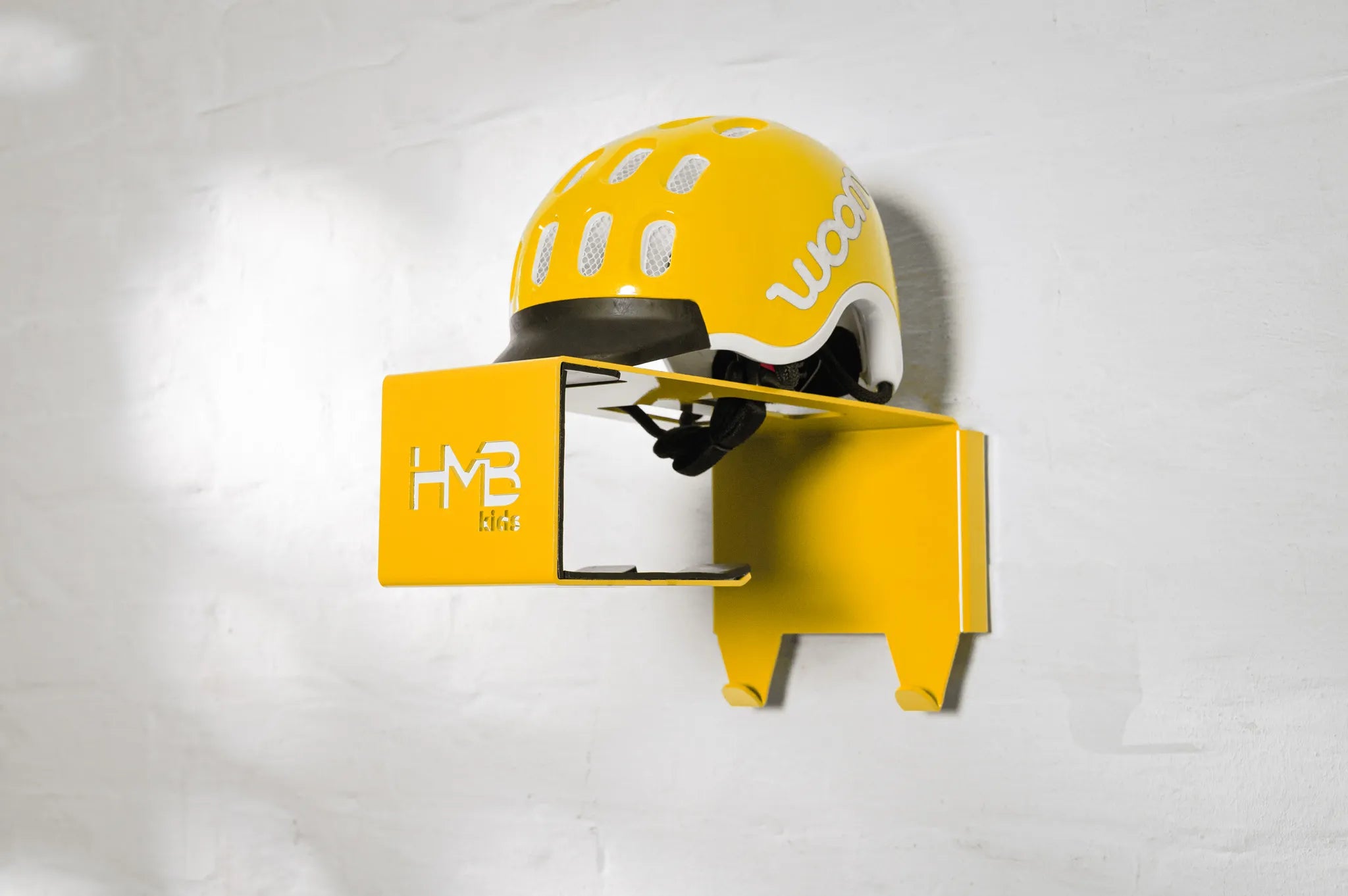 Perspective view of a yellow Hold My Bike (HMB) Kids wall mount with a matching yellow woom helmet on the top shelf, highlighting the laser-cut 'HMB Kids' branding and dual integrated equipment hooks.
