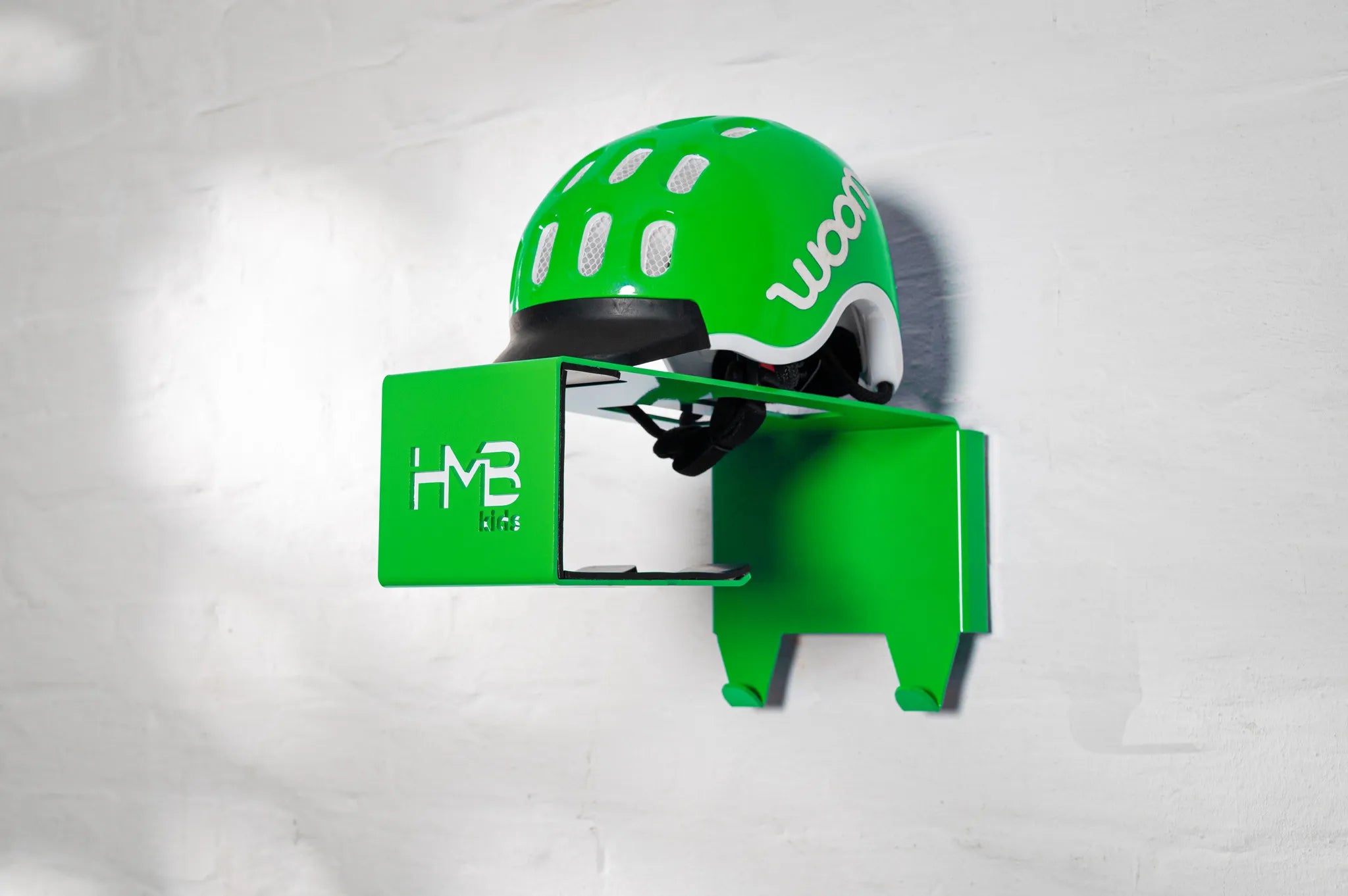 Perspective view of a green Hold My Bike (HMB) Kids wall mount with a matching green woom helmet on the top shelf, highlighting the laser-cut 'HMB Kids' branding and dual integrated equipment hooks.