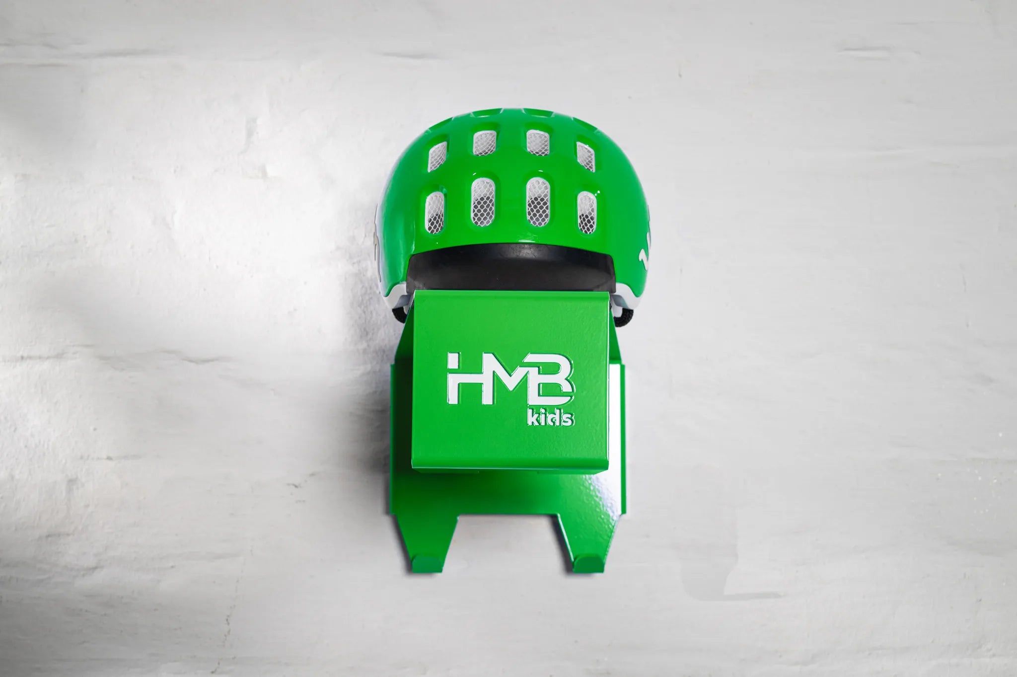 Front view of a green Hold My Bike (HMB) Kids wall mount with a matching green woom helmet resting on the top shelf, showcasing the laser-cut branding and two integrated hooks for cycling gear.