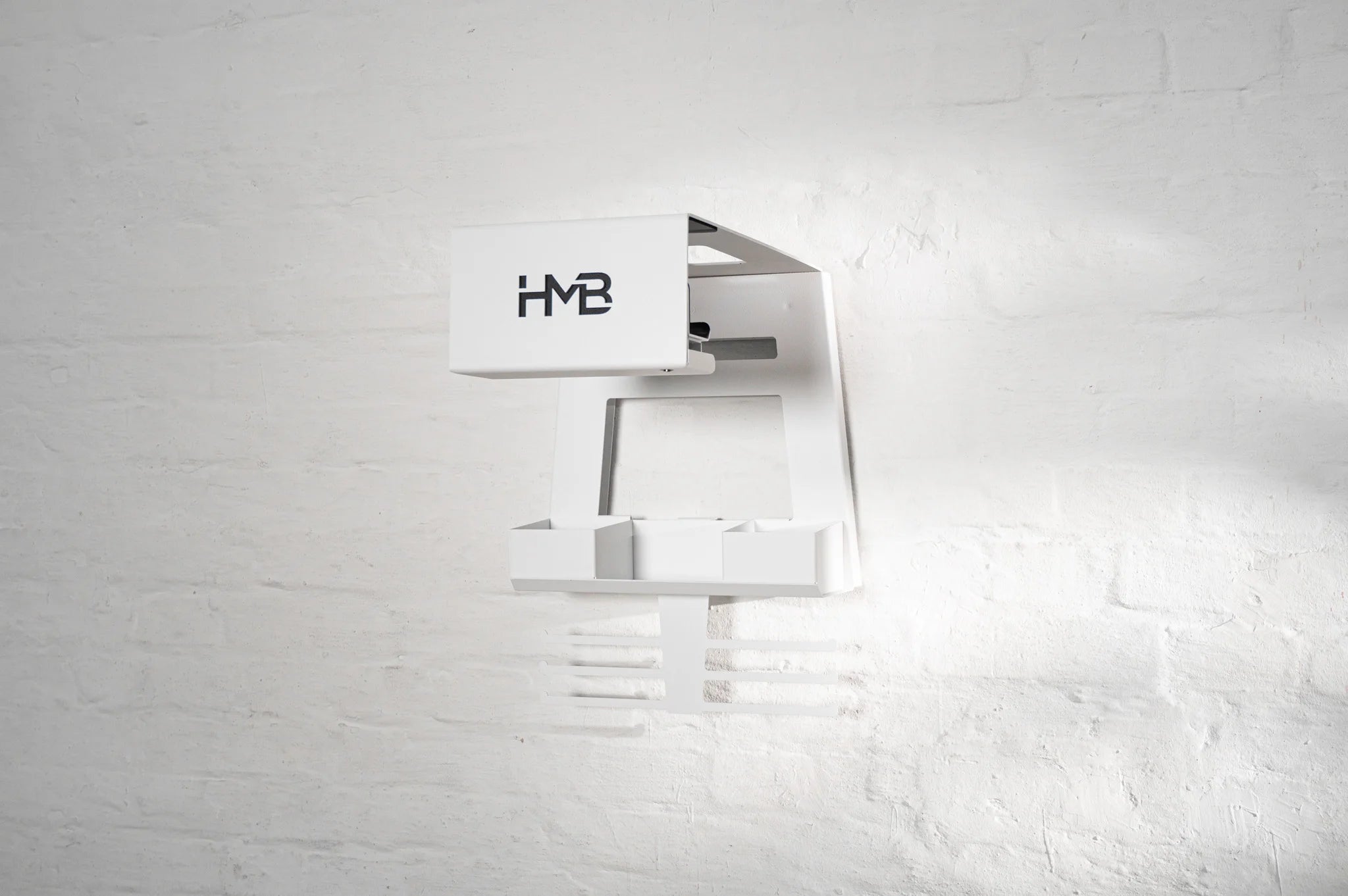 Perspective view of the white HMB Ultimate wall mount, highlighting the modular storage bins, integrated helmet shelf, and laser-cut 'HMB' branding against a minimalist white brick wall.