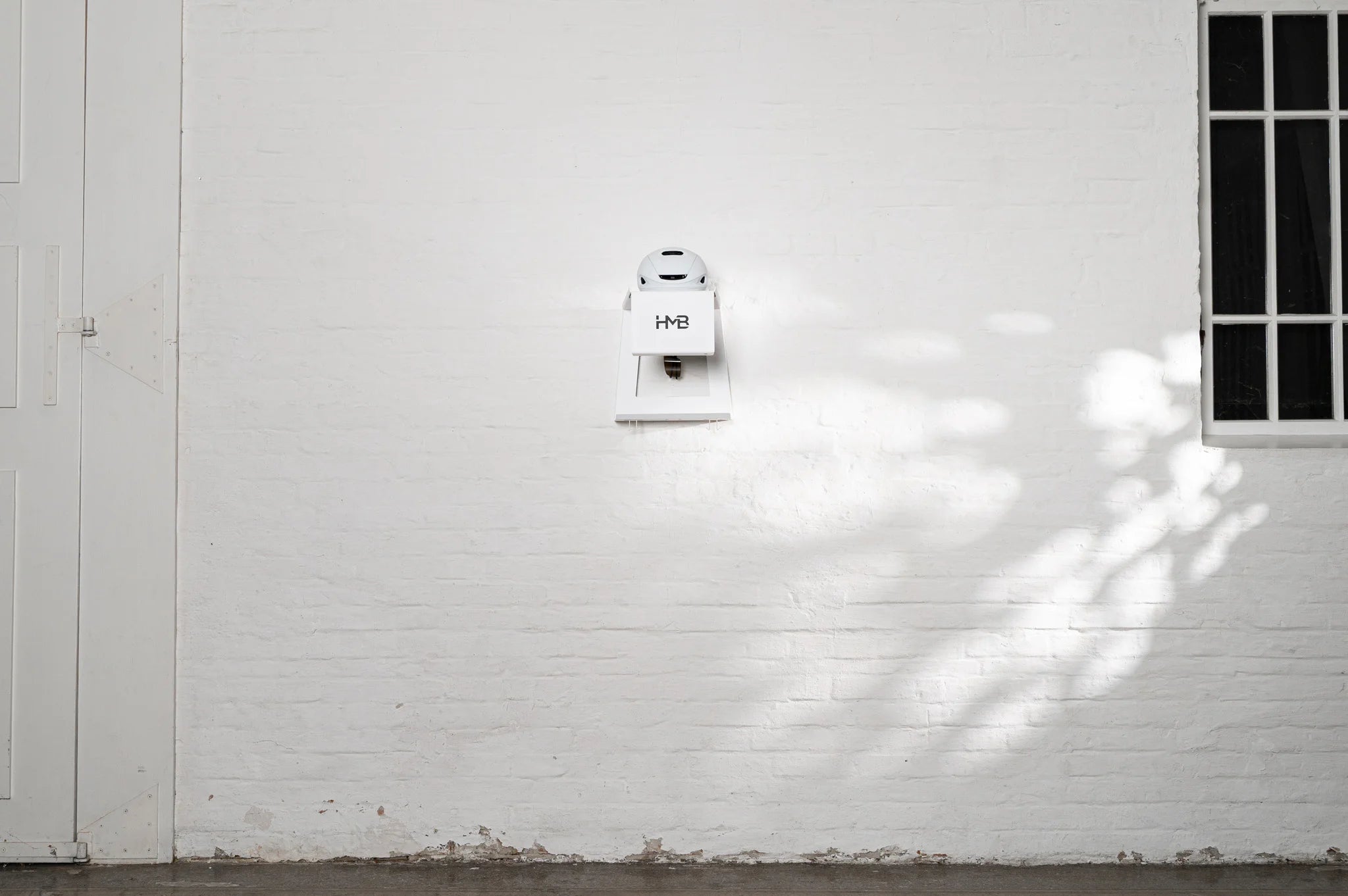 Wide architectural shot of a white HMB wall mount installed on a white brick wall, featuring a white cycling helmet on the top shelf and an integrated eyewear dock, showcasing its minimalist and clean design.