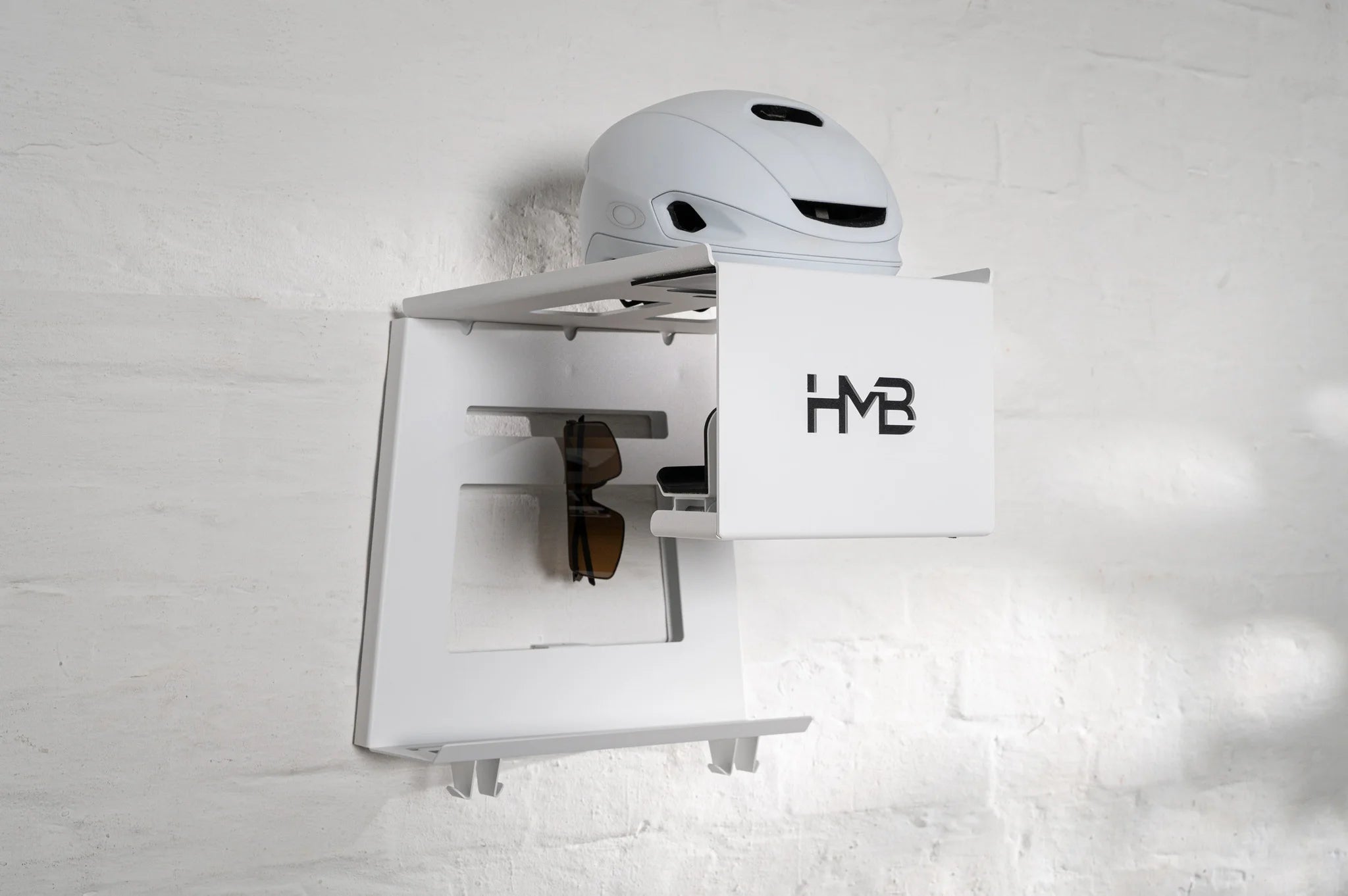 Perspective view of the white HMB Ultimate wall mount featuring a white cycling helmet on the top shelf, integrated eyewear storage for sunglasses, and a sturdy lower gear rack with two hooks for cycling apparel.