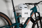 A teal Santa Cruz mountain bike stored on a white HMB Ultimate XL wall mount, featuring an integrated white helmet on the top shelf and an HMB wheel strap securing the front wheel for maximum stability