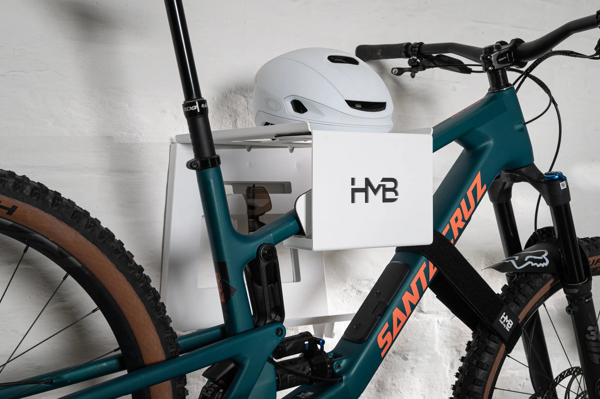A teal Santa Cruz mountain bike stored on a white HMB Ultimate XL wall mount, featuring an integrated white helmet on the top shelf and an HMB wheel strap securing the front wheel for maximum stability