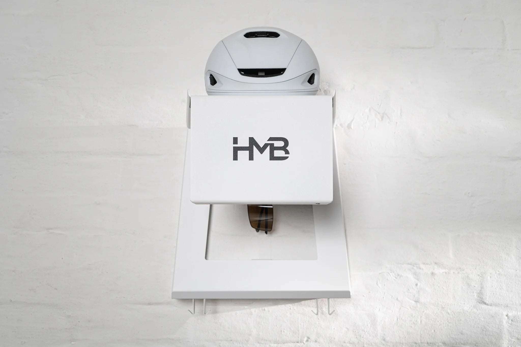 Front-facing view of the white HMB Ultimate wall mount installed on a white brick wall, featuring a white cycling helmet on the top shelf, integrated eyewear storage, and lower gear hooks.